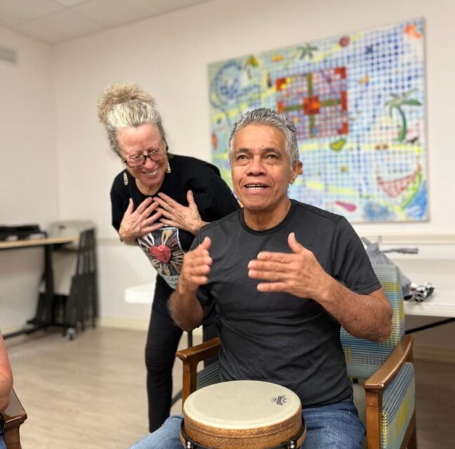 Lighthouse of Collier client playing a hand drum while a volunteer cheers him on
