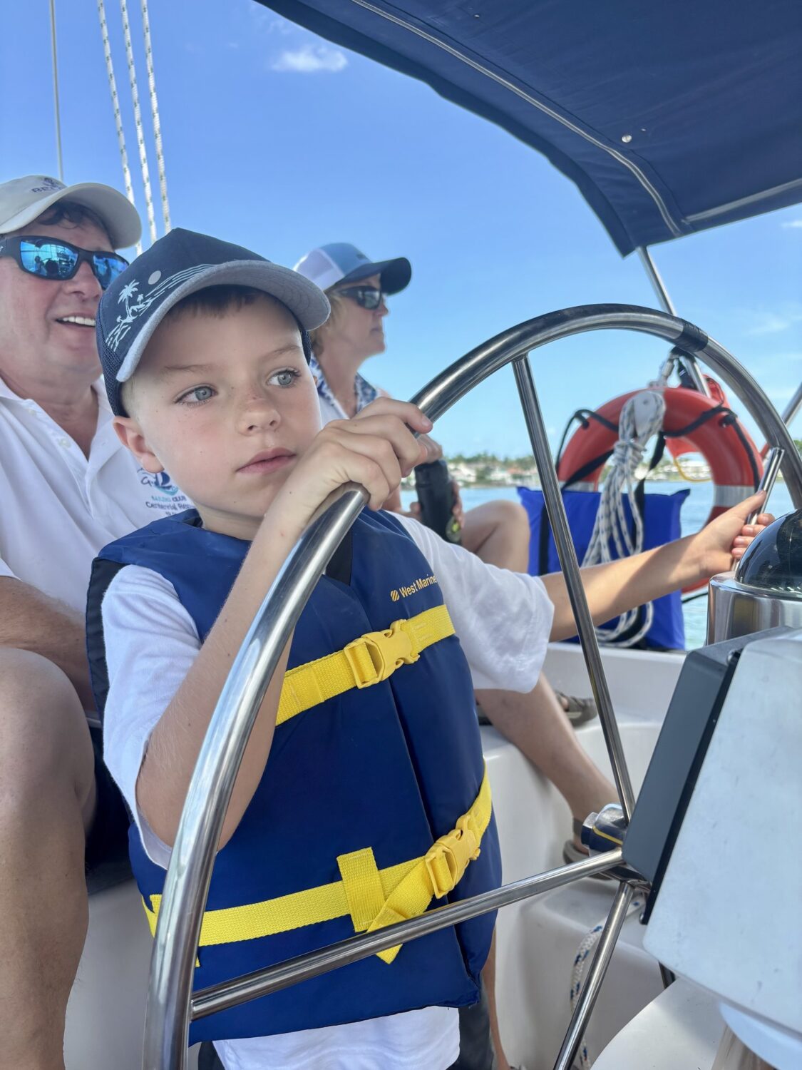 Young boy in a life vest steering a sailboat with volunteers alongside