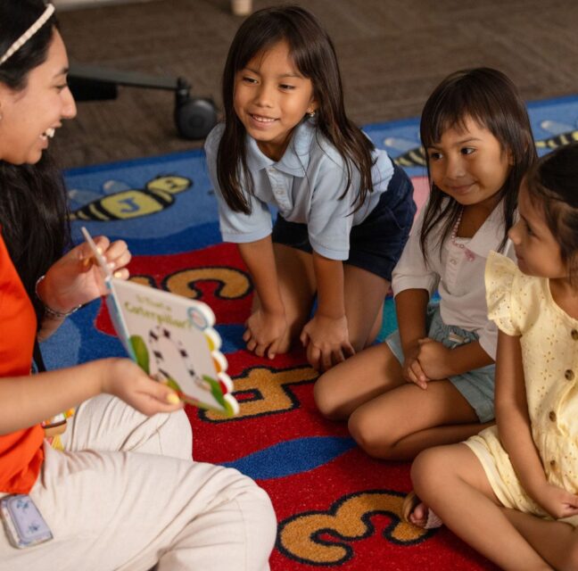 Teacher showing a lesson card to three young girls seated on a classroom rug