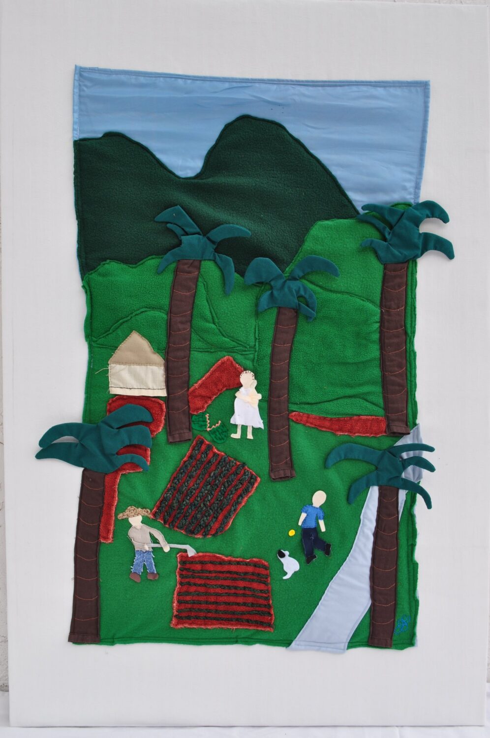 Green fabric artwork depicting a hillside scene with figures and houses